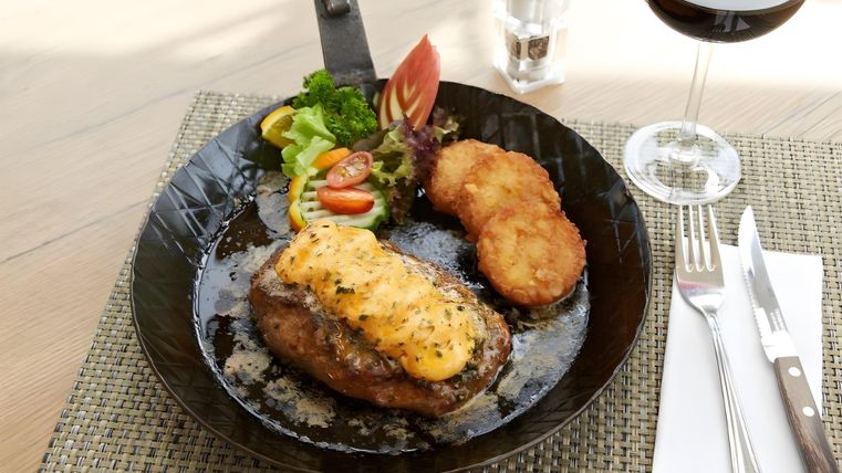 Ein saftiges Steak mit geschmolzenem Käse, begleitet von Beilagen aus frischem Salat, kleinen Tomaten und Kartoffelrösti. Dazu ein Glas Rotwein auf einem eleganten Tisch.