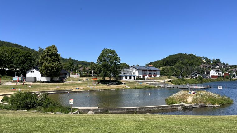 Ein ruhiger See umgeben von grünen Wiesen und Bäumen. Im Hintergrund sind einige Gebäude und Hügel zu sehen.