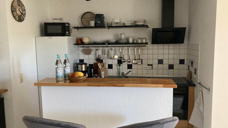 A modern kitchen with a wooden bar and two bar stools. Water bottles and fruit are on the countertop, while the shelves are well-organized.
