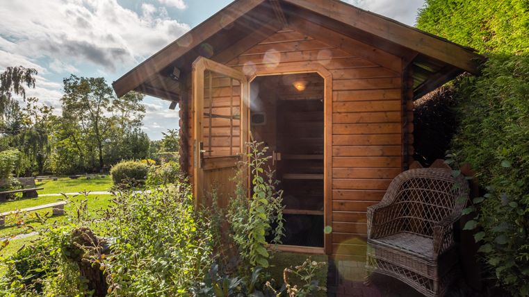 A small wooden sauna with an open door stands in a green garden. Next to the sauna is a cozy rattan bench.