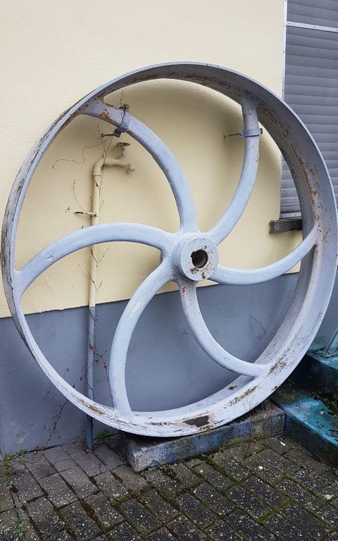 Ein großes, rostiges Rad steht an einer Wand. Der Hintergrund ist gelb und es sind einige Pflastersteine sichtbar.
