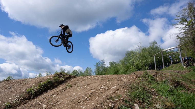 Een mountainbiker springt met zijn fiets over een heuvel. De lucht is blauw met enkele wolken en de omgeving is groen.