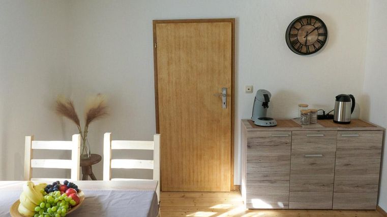 Eine helle Küche mit einem Tisch, auf dem eine Schale mit frischen Früchten steht. An der Wand befindet sich eine Uhr und eine verschlossene Tür.