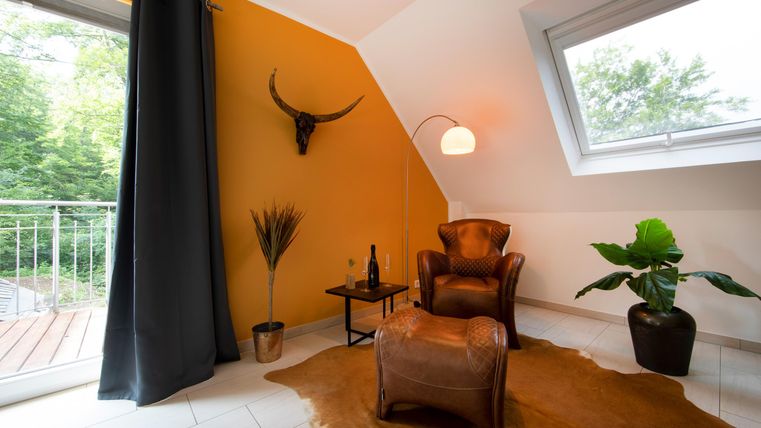 Ein gemütlicher Raum mit einer braunen Ledersessel und einem kleinen Tisch. Die Wände sind gelb gestrichen und es gibt Pflanzen sowie große Fenster, die viel Licht hereinlassen.