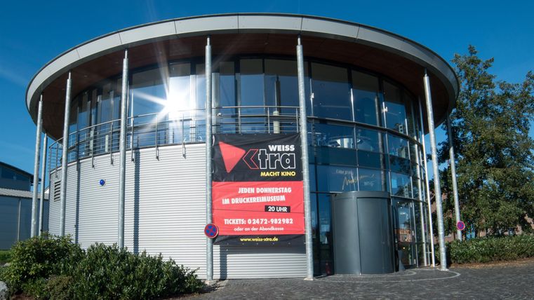 Ein modernes Gebäude mit großen Fenstern und einer runden Form. An der Fassade befindet sich ein auffälliges Schild mit Informationen.