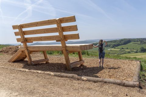 A huge wooden bench stands in a rural landscape. A person is standing next to it, looking into the distance.