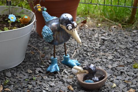 Keramikkunstwerk eines Vogels mit blauen Flügeln und großen Augen, steht auf Kies neben einem Blumentopf mit Blumenverzierung.