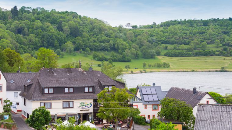 Ein gemütliches Restaurant am Ufer, umgeben von grünen Hügeln und sanften Landschaften. Im Vordergrund sind Tische im Freien und ein ruhiger Fluss zu sehen.