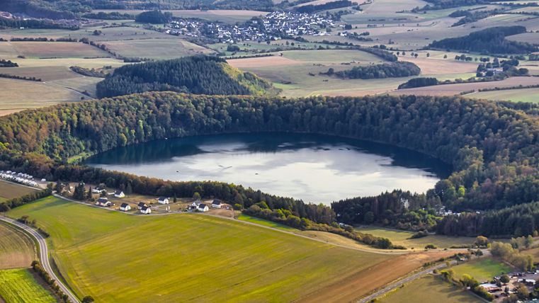 Ein malerischer See umgeben von Wäldern und Wiesen. Im Hintergrund sind sanfte Hügel und kleine Dörfer zu sehen.