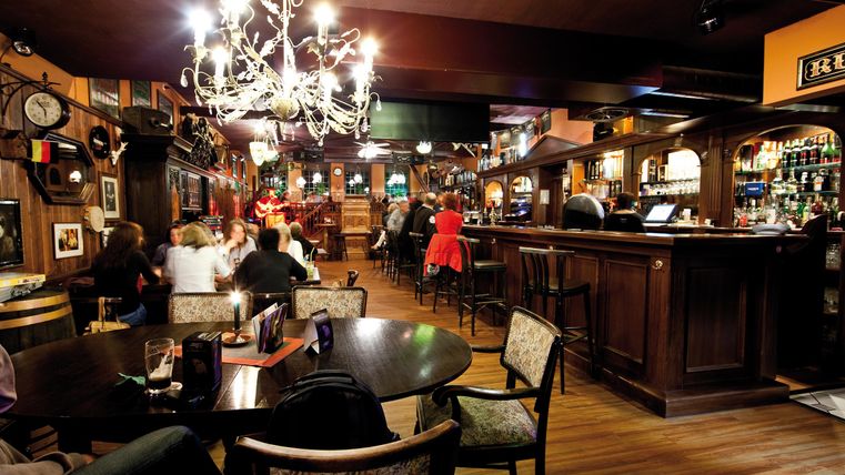 Eine gemütliche Bar mit Holzmöbeln und einer warmen Atmosphäre. Gäste genießen Getränke und Live-Musik im Hintergrund.