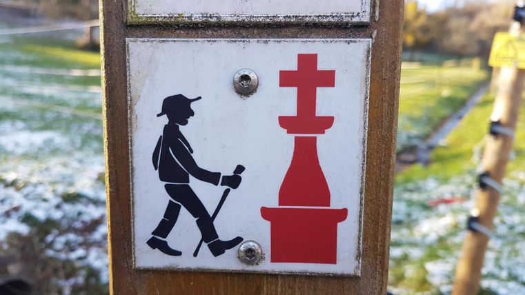 Ein Schild zeigt einen Wanderer mit Stock, der auf einen roten Wegweiser aufmerksam ist. Es weist auf einen Aussichtspunkt oder ein Ziel hin.