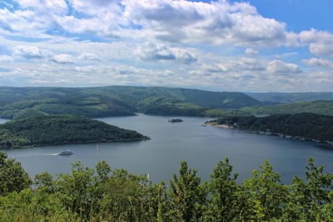 Eine malerische Landschaft mit einem See, umgeben von grünen Hügeln. Der Himmel ist blau mit einigen Wolken.