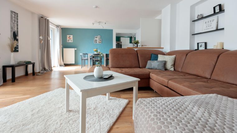 Ein modernes Wohnzimmer mit einem großen braunen Sofa, einem weißen Couchtisch und einem gemütlichen Teppich. Im Hintergrund sind eine blaue Wand und ein Esstisch sichtbar.