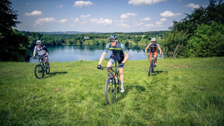 Three mountain bikers ride along a green hill overlooking a lake. The sky is blue and dotted with some clouds.