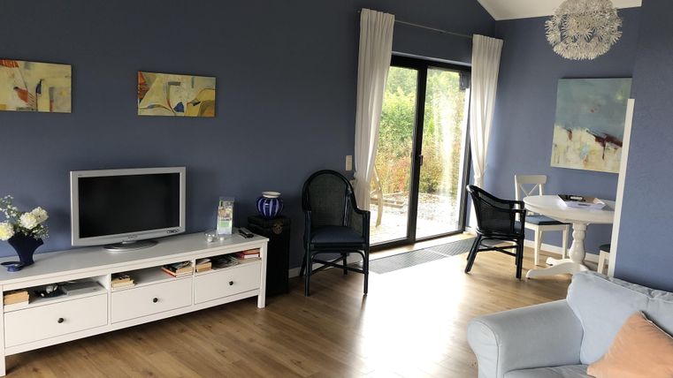 Een moderne woonkamer met blauwe muren en een houten vloer. Er is een tv-meubel, een gezellige fauteuil en een eettafel met stoelen.