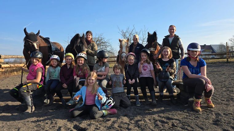 A group of children and adults stands and sits at a riding stable, surrounded by horses. The atmosphere is cheerful and sunny.