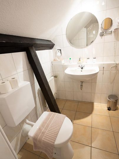 A small bathroom with a sink, a mirror, and a toilet. The walls are tiled with white tiles, and the floor is tile-like.