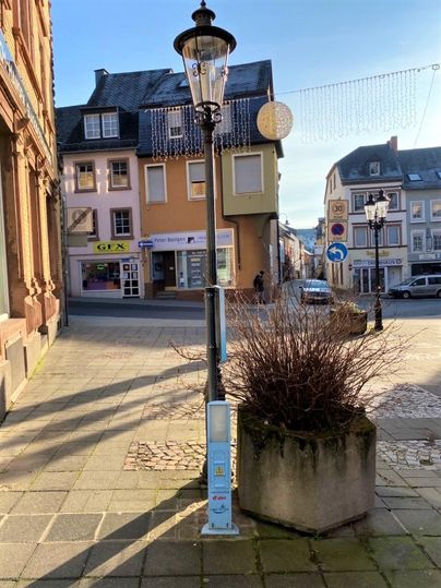 A sunny street scene with a street lamp and a plant pot. In the background, various buildings and a traffic road can be seen.
