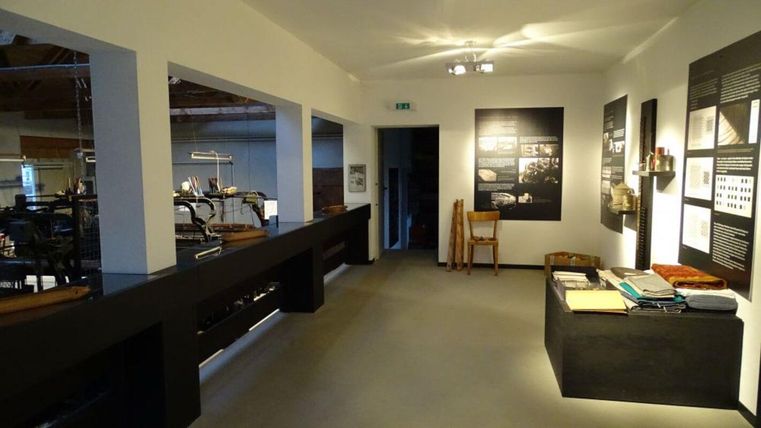 Ein moderner Ausstellungsraum mit hellen Wänden und Informationen an den Wänden. Im Raum sind verschiedene Tische und Stühle platziert.