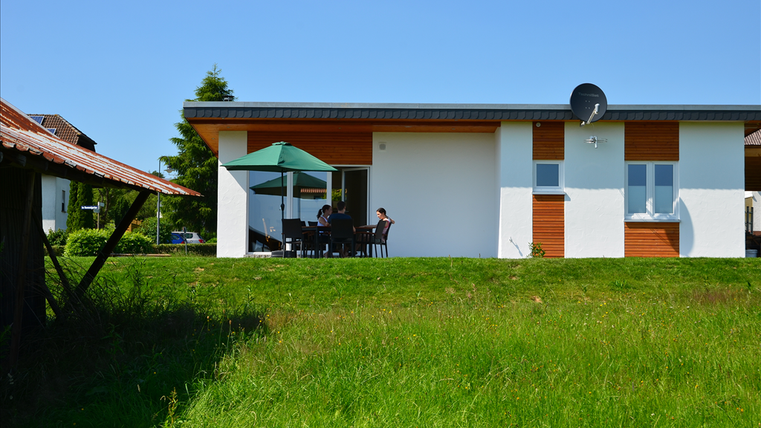 A modern house with a terrace and a green lawn. People are sitting on the terrace under a sunshade.
