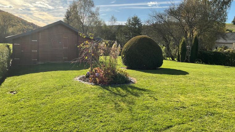 A well-kept garden with green grass, colorful flowers, and a round bush. In the background, there is a small wooden gazebo.