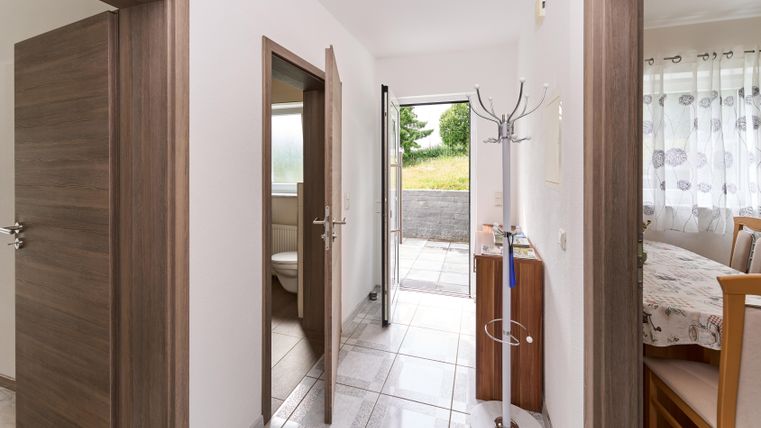 Ein heller Flur mit geöffneten Türen und einem Blick ins Freie. An der Wand steht ein Kleiderständer und ein Tisch ist im Hintergrund sichtbar.