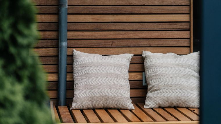 Ein Holzbank mit zwei gemütlichen Kissen steht an einer Holzwand. Die Umgebung wirkt warm und einladend.