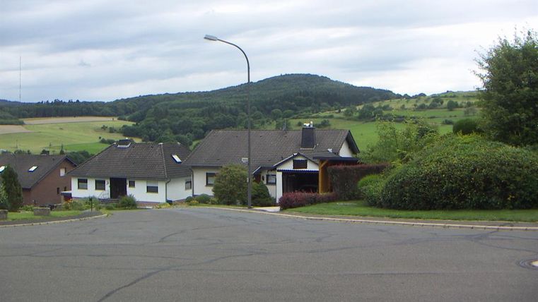 A quiet residential area with several houses and a gentle hill backdrop in the background. The street is wide and well-maintained, surrounded by green spaces.