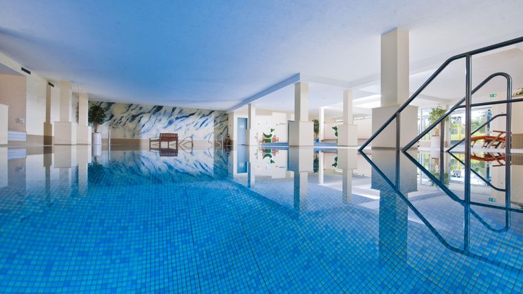 Ein modernes Hallenbad mit blauem Wasser und glitzernden Fliesen. Die Umgebung ist hell und einladend, mit Pflanzen und einem entspannenden Ambiente.