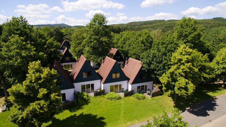 A group of stylish houses with red roofs surrounded by lush greenery. In the background, gentle hills stretch beneath a blue sky.