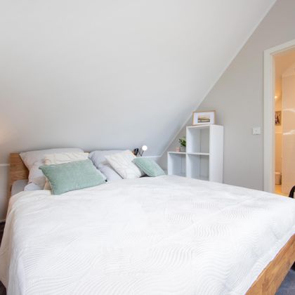 Een lichte slaapkamer met een groot bed en gezellige kussens. Op de achtergrond is een moderne badkamer met douche zichtbaar.