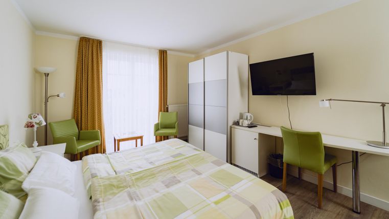 Ein helles Hotelzimmer mit einem Doppelbett, einem Schreibtisch und zwei Stühlen. Der Raum hat große Fenster und eine moderne Einrichtung.