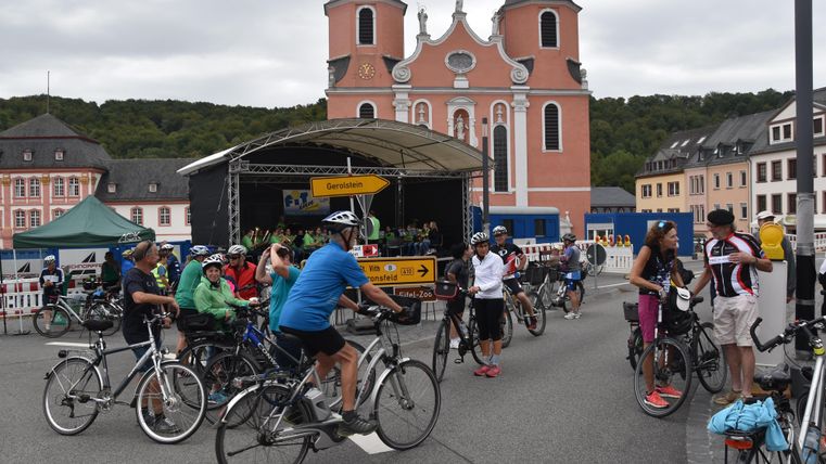 Eine Gruppe von Radfahrern versammelt sich vor einer historischen Kirche. Im Hintergrund sind Gebäude und eine Bühne zu sehen.