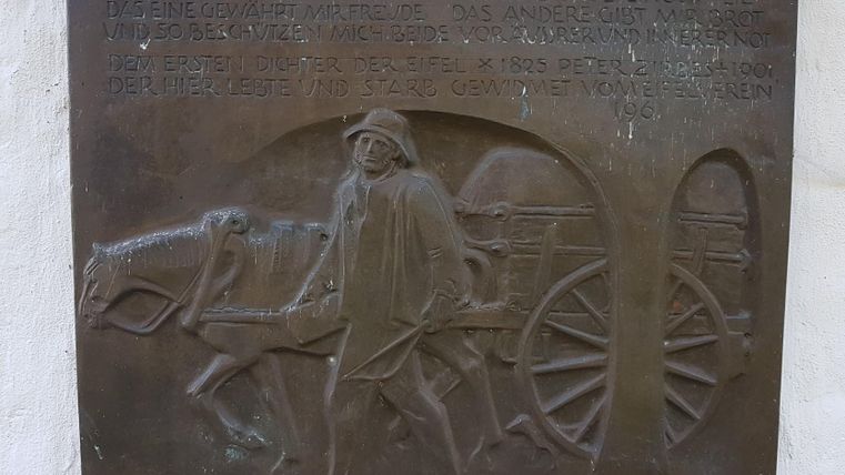 Eine bronzene Reliefplatte zeigt einen Mann, der ein Pferd zieht und eine Wagen. Der Hintergrund enthält Text in einer alten Schrift.