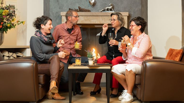 Eine gemütliche Runde von vier Personen, die Getränke genießen und lachen. Sie sitzen in einem Wohnzimmer vor einem Kamin.