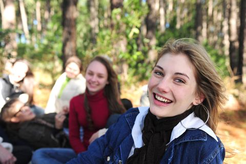 Eine Gruppe junger Menschen sitzt im Wald und lacht freundlich. Eine Frau mit blonden Haaren lächelt in die Kamera.