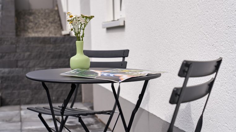 Ein kleiner Tisch mit zwei Stühlen steht an einer Wand. Auf dem Tisch steht eine grüne Vase mit Blumen und eine Zeitschrift liegt darauf.