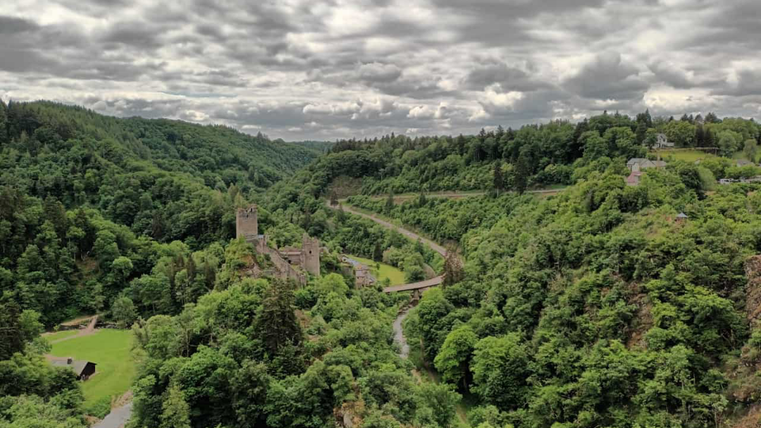 Een indrukwekkend landschap met groene heuvels en een oude kasteelruïne. De lucht is bewolkt en de omgeving is omringd door bomen.
