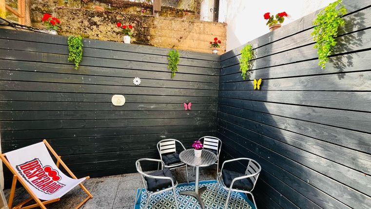 A cozy courtyard with black wall cladding and green plants. Two chairs and a table are ready to enjoy the relaxed atmosphere.