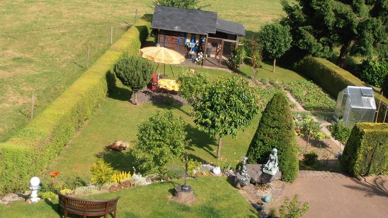 Ein schöner Garten mit üppigem Grün und verschiedenen Pflanzen. Im Hintergrund steht ein Gartenhaus und eine Terrasse mit einem Sonnenschirm.