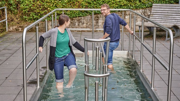 Zwei Personen gehen in einem Wassertretbecken. Sie lachen und scheinen die frische, nasse Erfahrung zu genießen.