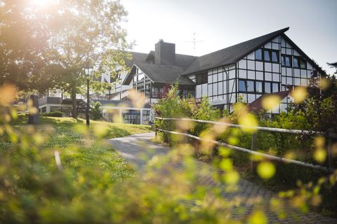 Ein modernes Gebäude mit einem traditionellen Fachwerkstil, umgeben von üppiger Vegetation. Der Weg führt sanft zu der Unterkunft unter einem klaren Himmel.