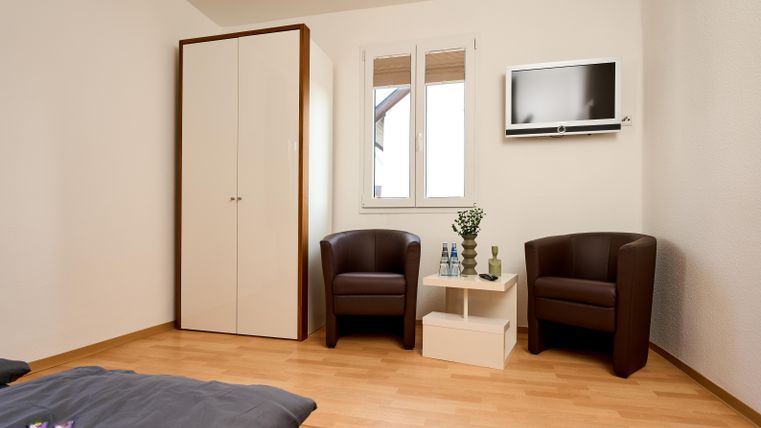 Ein einfaches und modernes Zimmer mit einem Doppelbett, zwei bequemen Stühlen und einem kleinen Tisch. An der Wand hängt ein Fernseher und es gibt einen Schrank sowie Fenster für Tageslichteinfall.