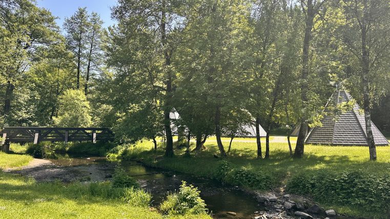 Eine ruhige Landschaft mit einem kleinen Fluss und Brücke. Im Hintergrund sind Bäume und pyramidenförmige Gebäude zu sehen.