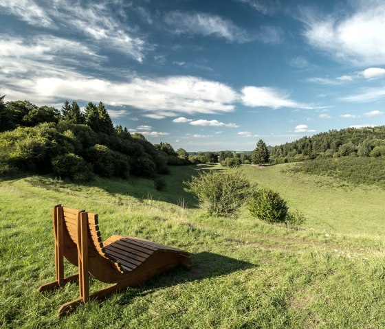 Houten ligstoel op een weiland met uitzicht op de groene Papenkaule en een blauwe lucht met wolken., © Eifel Tourismus GmbH, Dominik Ketz