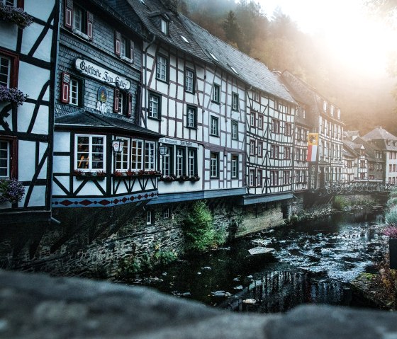 Fachwerkhäuser in Monschau am Fluss Rur, beleuchtet vom Sonnenaufgang. Herbstliche Bäume und eine ruhige Atmosphäre prägen das Bild., © Tourismus NRW e.V.