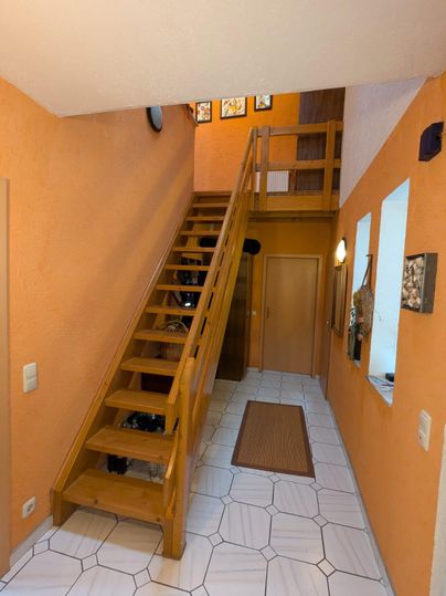 A bright hallway with orange walls and a wooden staircase. The floor is designed with white tiles and a carpet in the middle.