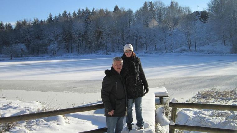 A snowy landscape picture with a frozen lake. Two people are standing on a wooden plank in front of the lake.