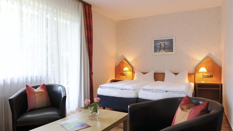 Ein gemütliches Hotelzimmer mit zwei Betten und sanften Farben. Es gibt zwei Sessel und einen kleinen Tisch, umgeben von natürlichem Licht.