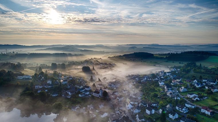 Eine malerische Landschaft mit einem Dorf in der Morgendämmerung. Der Himmel ist klar mit sanften Wolken und Nebel, der über die Felder zieht.